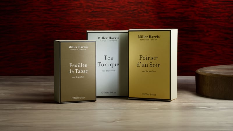 Three Miller Harris fragrance boxes on a light wood counter with a red wood background